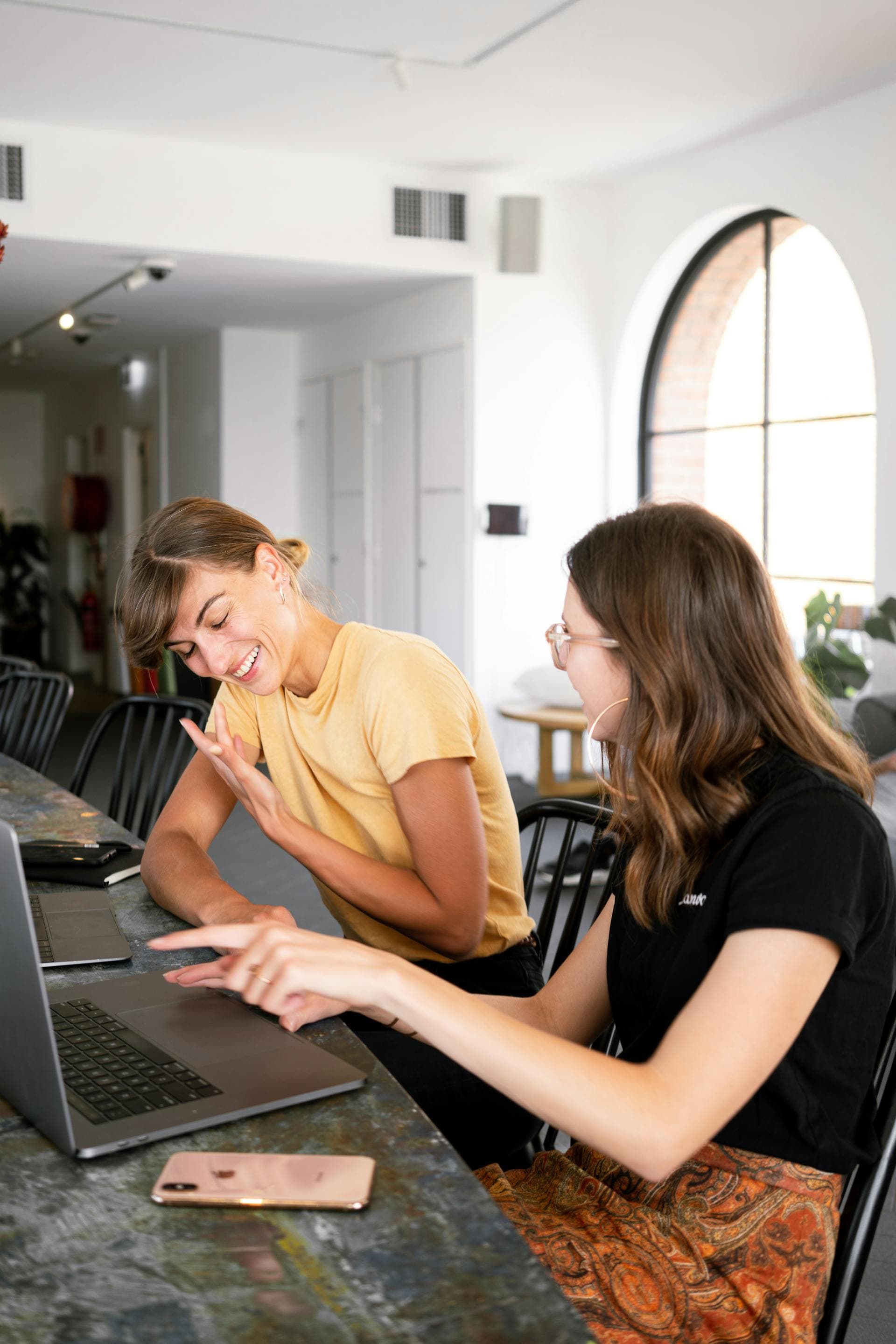 Two women collaborating over a laptop, laughing and sharing ideas in a bright workspace